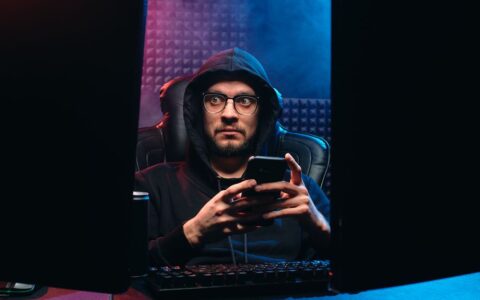 Man in hoodie with eyeglasses using smartphone between computer screens in a dark room.