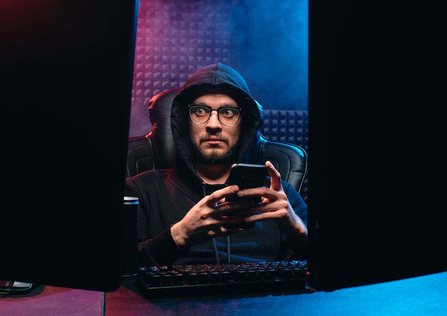 Man in hoodie with eyeglasses using smartphone between computer screens in a dark room.